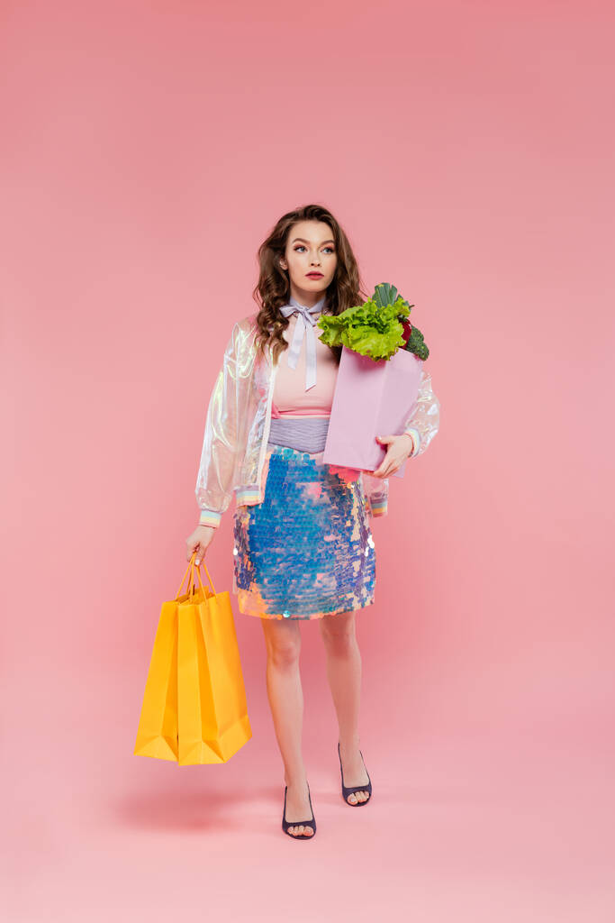 piękna młoda kobieta niosąca torby na zakupy i torbę spożywczą, stojąca na różowym tle, fotografia koncepcyjna, obowiązki domowe, jedzenie i warzywa, koncepcja gospodyni domowej  - Zdjęcie, obraz