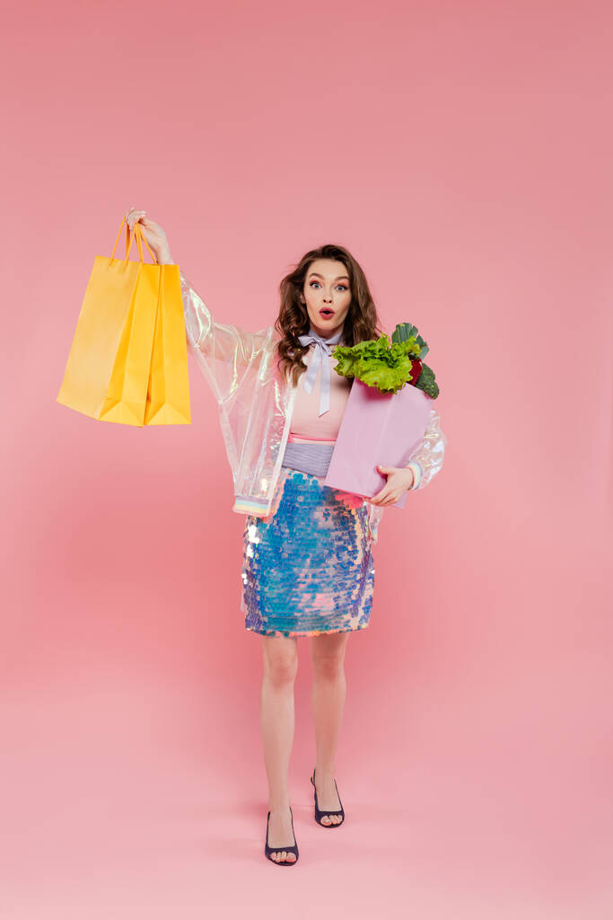 amazed young woman carrying shopping bags and grocery bag with vegetables, standing on pink background, conceptual photography, home duties, housewife concept  - Photo, Image
