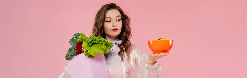 housewife concept, attractive young woman carrying grocery bag with vegetables and bowl with corn flakes, model with wavy hair on pink background, conceptual photography, home duties, banner - Photo, Image