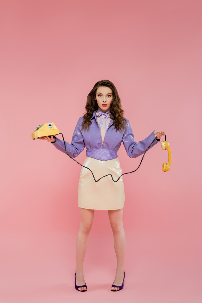 doll pose, young woman with wavy hair holding handset and retro phone, trendy outfit, brunette model in purple jacket posing and looking at camera on pink background, studio shot  - Photo, Image