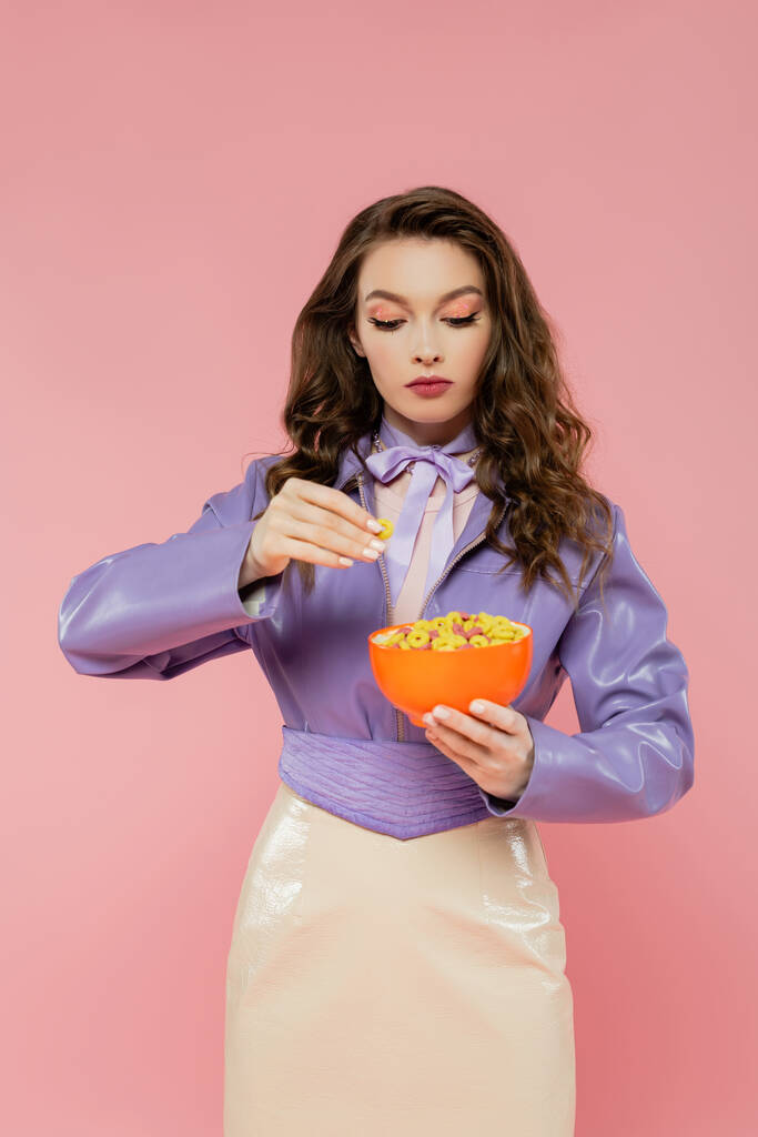 Konzeptfotografie, brünette Frau mit welligem Haar, die vorgibt, eine Puppe zu sein, Schale mit Cornflakes in der Hand, beim Frühstück, posiert auf rosa Hintergrund, trendige lila Jacke - Foto, Bild