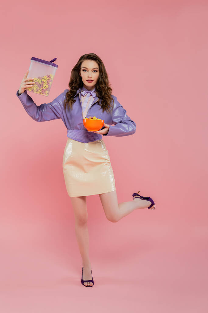 концептуальная фотография, брюнетка с волнистыми волосами, притворяющаяся куклой, стоя на одной ноге, держа контейнер с кукурузными хлопьями, завтрак, позируя на розовом фоне, стильная фиолетовая куртка - Фото, изображение