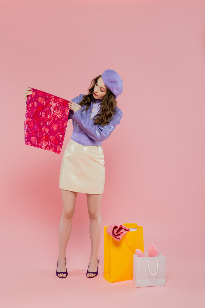 Mode und Konsum, attraktive junge Frau in Baskenmütze, die lebendige farbige Kleidung in der Nähe von Einkaufstaschen auf rosa Hintergrund hält, Konsumverhalten, in trendiger Lederjacke und Rock stehend, volle Länge - Foto, Bild