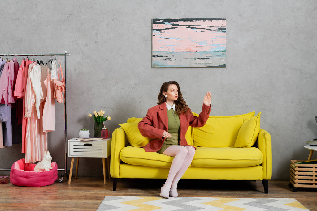 Konzeptfotografie, Frau wie eine Puppe auf gelbem Sofa sitzend, unnatürlich gestikulierend im modernen Wohnzimmer, gut gekleidet und schön, moderne Hauseinrichtung, Rollenspiel, Puppenleben  - Foto, Bild