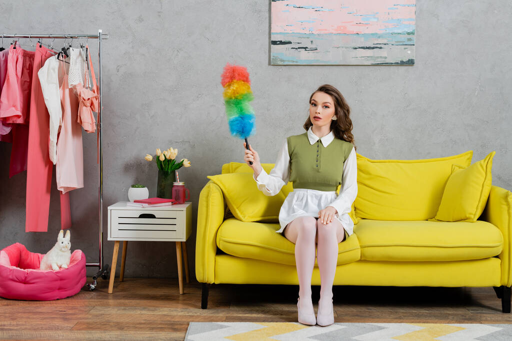 Hauswirtschaftskonzept, junge Frau mit welligem Haar auf Couch sitzend und Staubbürste in der Hand, Hausfrau im Kleid und weißen Strumpfhosen vor der Kamera, häusliches Leben, posierend wie eine Puppe  - Foto, Bild