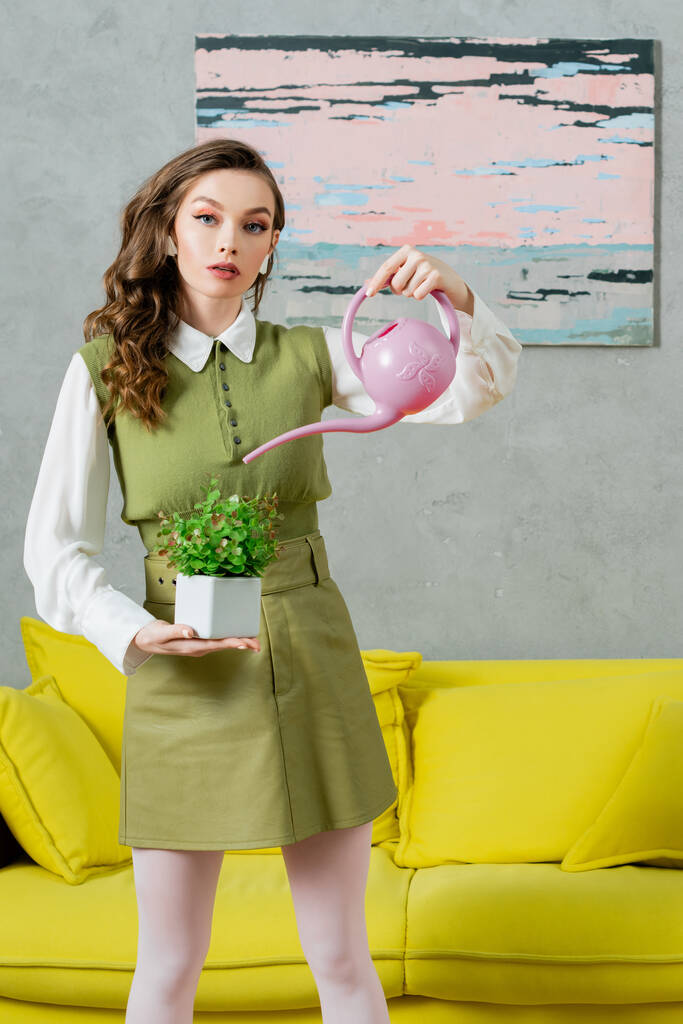 eco-friendly, conceito de limpeza, jovem com cabelo ondulado regando planta verde, crescimento e botânica, limpeza, bela dona de casa olhando para a câmera e de pé na sala de estar  - Foto, Imagem