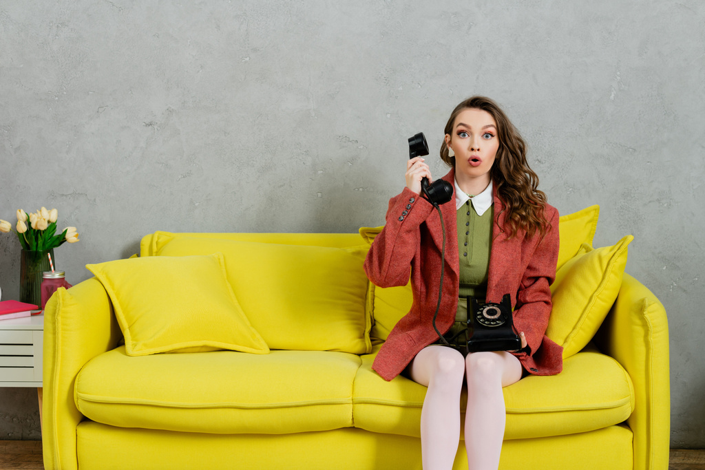 τηλέφωνο, έκπληκτη γυναίκα με κυματιστά μαλλιά κάθεται σε κίτρινο καναπέ, νοικοκυρά μιλάμε σε ρετρό τηλέφωνο, ενεργεί σαν μια κούκλα, κοιτάζοντας κάμερα με ανοιχτό το στόμα, σοκ, γκρι τοίχο  - Φωτογραφία, εικόνα