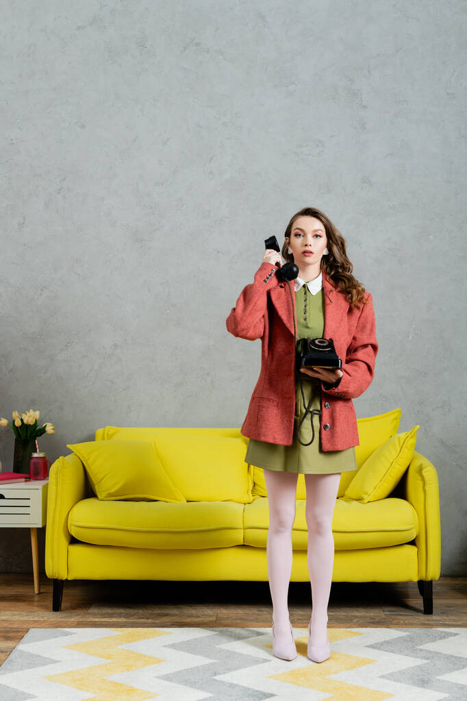 vintage vibes, beautiful woman with brunette and wavy hair standing near yellow couch, housewife talking on retro telephone, posing like a doll, looking at camera, modern interior, living room  - Photo, Image