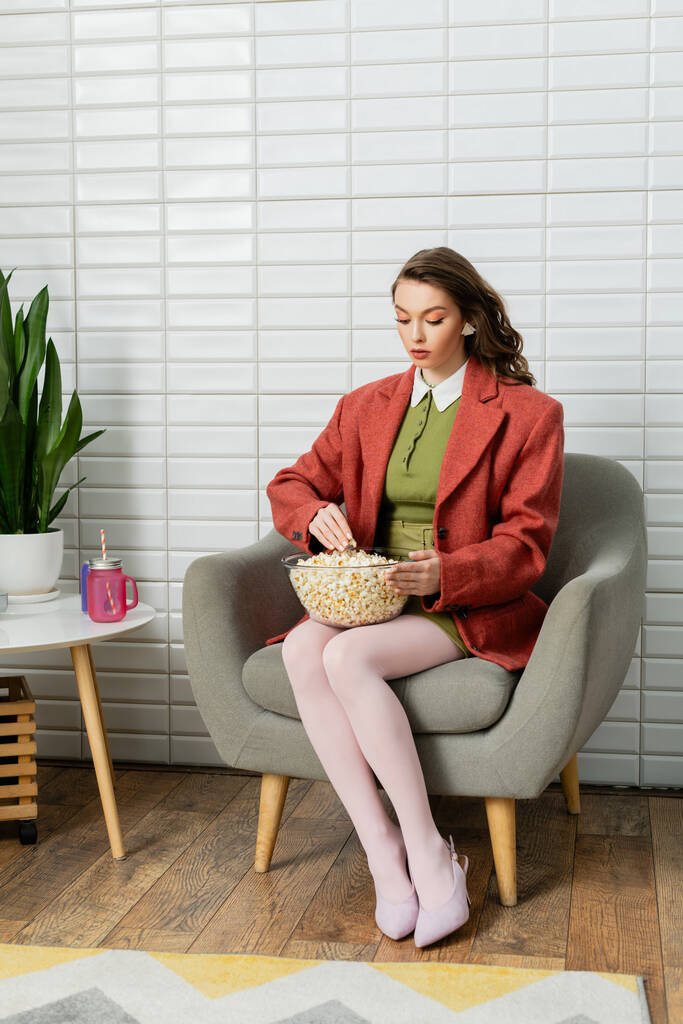fotografia koncepcyjna, młoda kobieta z falowanymi włosami brunetki siedząca na wygodnym fotelu, zachowująca się jak lalka, patrząca na miskę z popcornem, rozrywka, domowa rozrywka, przekąska filmowa  - Zdjęcie, obraz