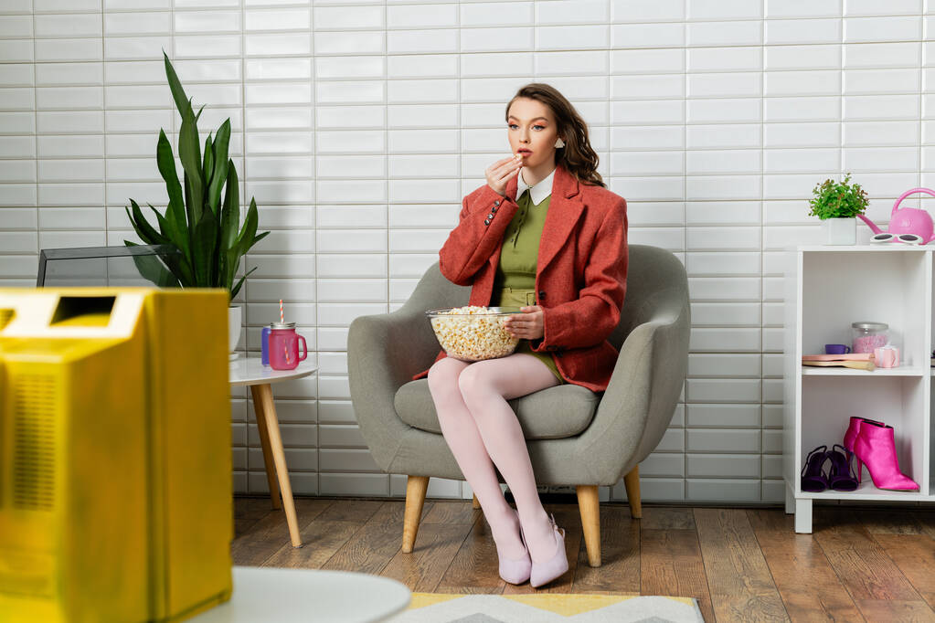 концептуальная фотография, молодая женщина с волосами брюнетки, сидящая на удобном кресле, смотрящая телевизор и питающаяся кукурузой, отдых, домашние развлечения, закуска в кино, Doll like  - Фото, изображение