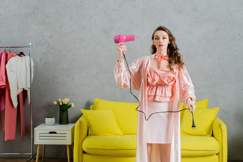 fotografía conceptual, mujer actuando como una muñeca, la vida doméstica, ama de casa en bata de seda rosa sosteniendo secador de pelo y enchufe, de pie cerca de entrenador amarillo en la sala de estar moderna, fingiendo como secar el cabello  - Foto, imagen