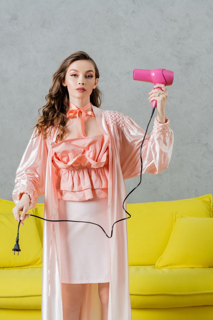 концептуальна фотографія, жінка діє як лялька, красива домогосподарка в рожевому шовковому халаті, що тримає фен і вилка, стоїть біля жовтого тренера в сучасній вітальні, прикидаючись сухим волоссям  - Фото, зображення