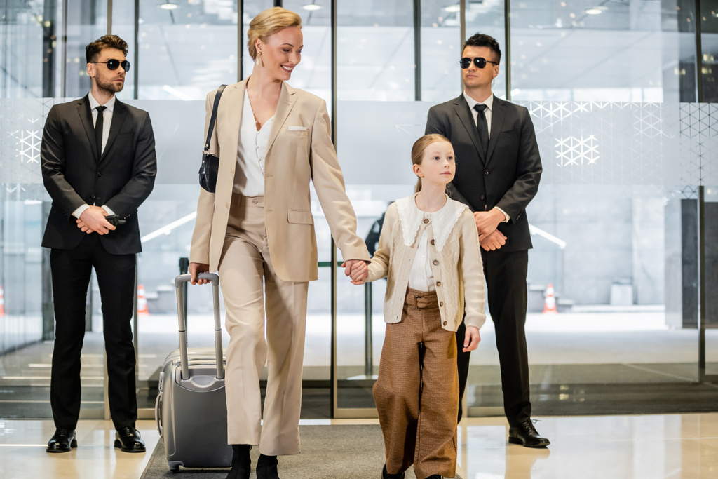 osobní bezpečnostní služba, dva bodyguardi ve formálním oblečení a slunečních brýlích stojící u vchodu do hotelu, šťastná matka a dítě držící se za ruce a kráčející se zavazadly, vstup do lobby, luxusní životní styl  - Fotografie, Obrázek
