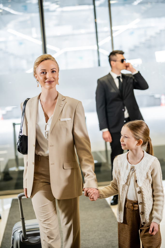 luxury lifestyle, rich life, personal security, cheerful woman with child, blonde woman holding hands with girl and walking inside of hotel, bodyguard in suit standing on blurred background  - Photo, Image