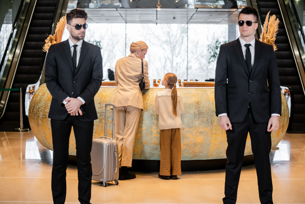 personal security, luxury lifestyle, blonde mother with child standing at reception desk, woman and preteen girl during check in, bodyguards in suit and sunglasses protecting rich family in hotel  - Photo, Image