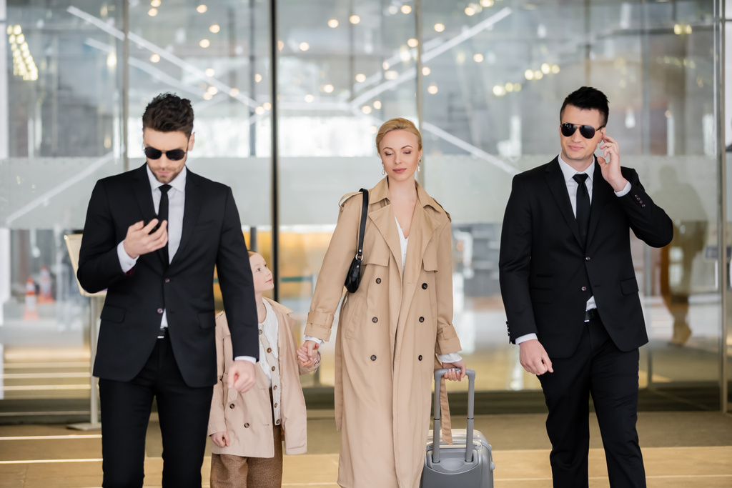 personal security concept, two bodyguards in suits and sunglasses protecting blonde woman with child with luggage entering hotel, wearing autumnal clothes, luxury and rich lifestyle  - Photo, Image