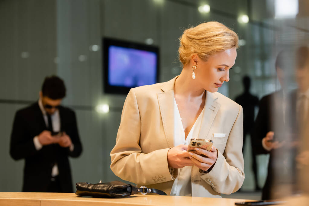 personal security, blonde woman in suit holding smartphone near reception desk, two bodyguards standing on blurred background, digital age, hospitality industry, personal security  - Photo, Image