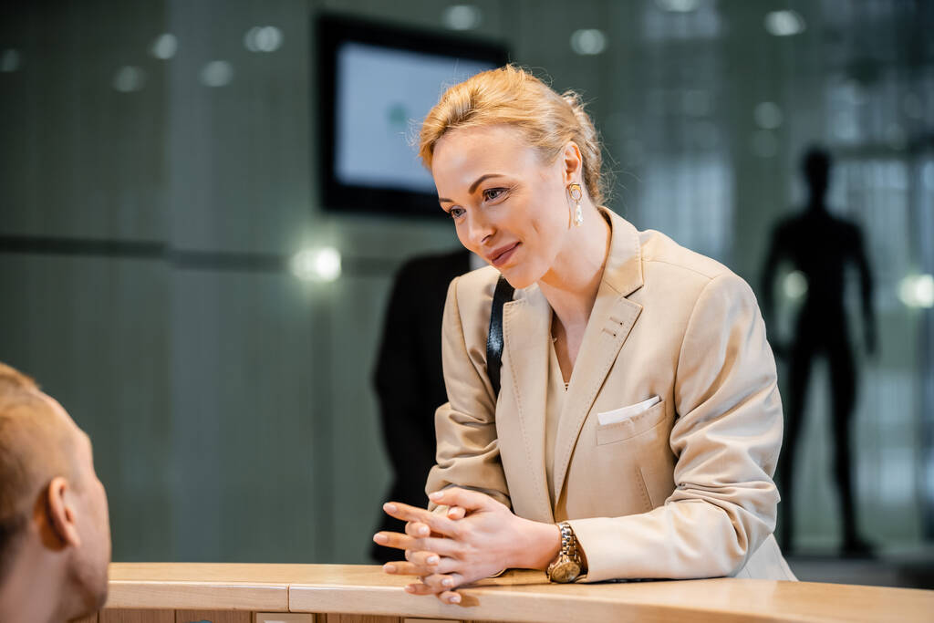 polite businesswoman talking to receptionist in hotel, hospitality industry, blonde and cheerful woman communicating with hotel staff, personal security, private safety, bodyguards on background  - Photo, Image