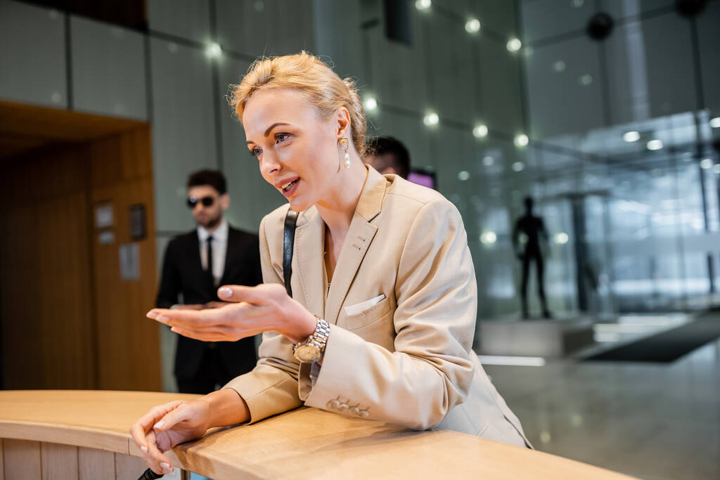 blonde woman in suit gesturing while talking at reception desk, personal security service concept, bodyguards in suits standing on blurred background, hotel industry, safety during travel  - Photo, Image