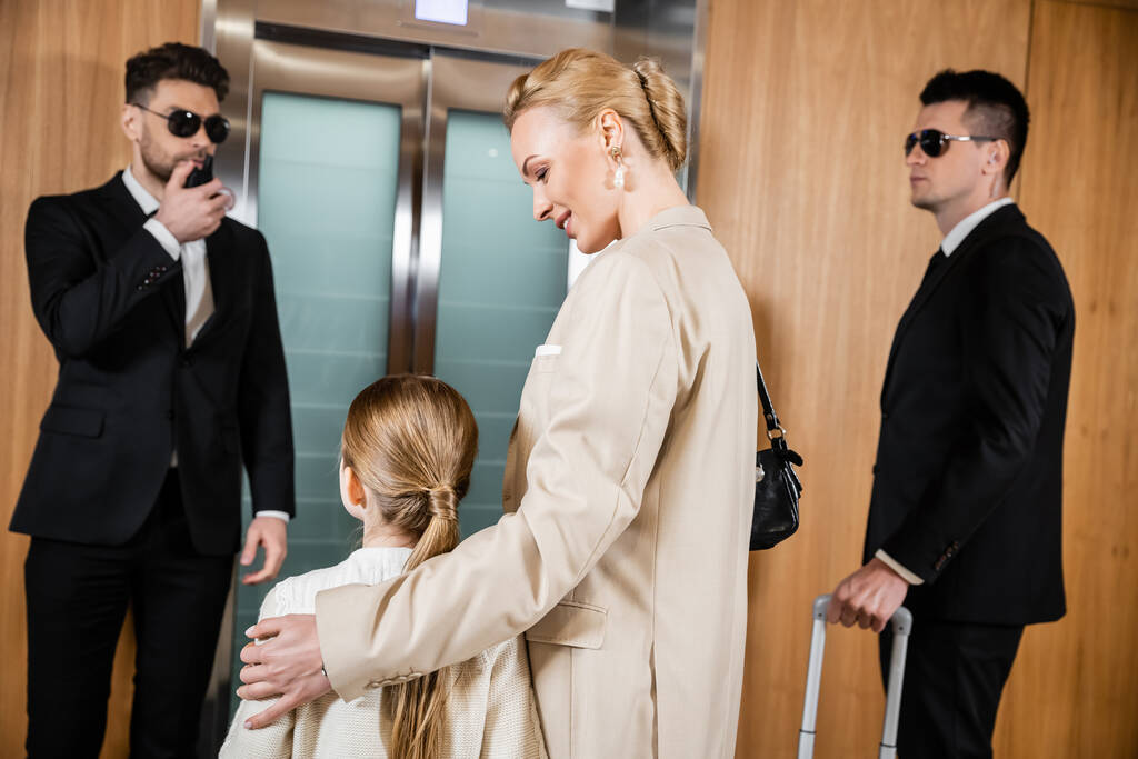 Блондинка мать обнимает ребенка, стоя рядом с лифтом отеля и телохранителей в костюмах, личной безопасности и защиты, успешной женщины и дочери подростка, семейных путешествий, службы безопасности  - Фото, изображение