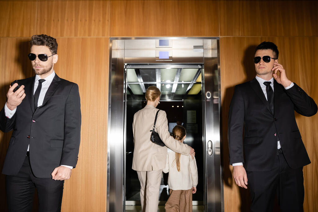 Özel güvenlik, anne ve kızı lüks otelin asansörüne biniyor, güvenliklerini koruyan iki koruma, takım elbiseli ve güneş gözlüklü yakışıklı adamlar kişisel güvenlik hizmetinde çalışıyor.  - Fotoğraf, Görsel