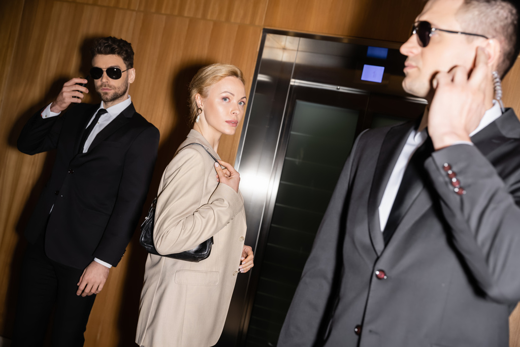 προσωπική ασφάλεια και προστασία έννοια, επιτυχημένη ξανθιά γυναίκα με τσάντα στέκεται κοντά ασανσέρ δίπλα σε σωματοφύλακες σε κοστούμια και γυαλιά ηλίου, πολυτελές ξενοδοχείο, θηλυκό επισκέπτη  - Φωτογραφία, εικόνα