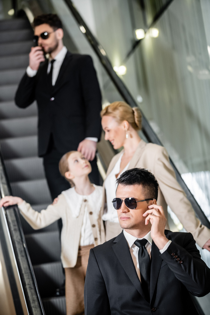 personal security concept, handsome bodyguards communicating while protecting safety of female clients, rich lifestyle, successful woman and preteen child standing on escalator of hotel - Photo, Image