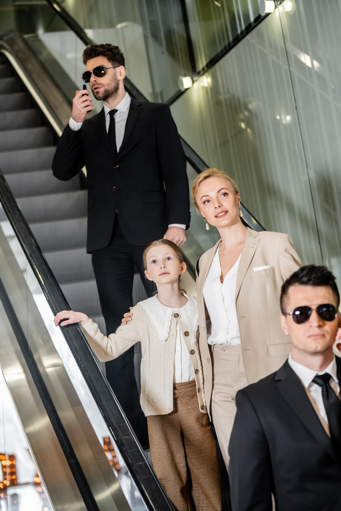концепція особистої безпеки, успішна жінка та її дочка стоять на ескалаторі розкішного готелю, двоє охоронців спілкуються, захищаючи безпеку клієнтів, багатий спосіб життя  - Фото, зображення