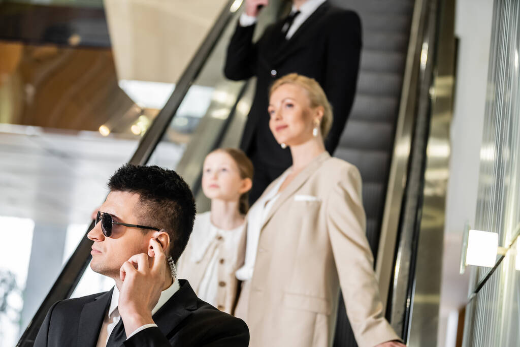 personal security concept, bodyguard communicating through earpiece while protecting safety of clients, rich lifestyle, successful woman and preteen daughter standing on escalator of hotel  - Photo, Image
