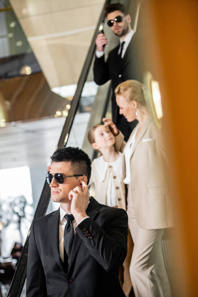 personal security concept, bodyguards communicating while protecting safety of clients, rich lifestyle, blonde and successful woman and preteen daughter standing on escalator of luxurious hotel  - Photo, Image