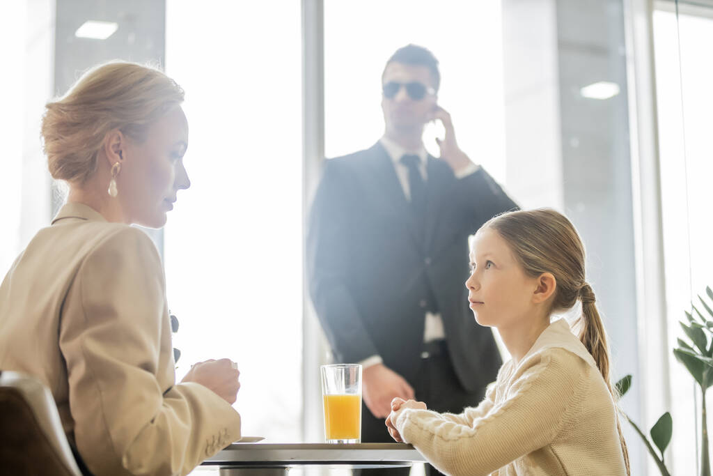 девочка-подросток разговаривает с матерью-блондинкой возле телохранителя на размытом фоне, служба безопасности, частная концепция безопасности, стакан апельсинового сока, личная безопасность  - Фото, изображение