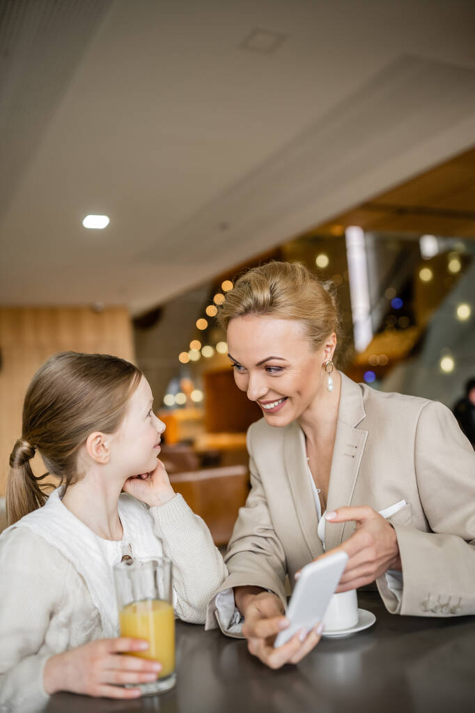 happy mother and daughter spending quality time together, blonde woman holding smartphone near daughter, digital age, working parent and kid, modern parenting, family bonding  - Photo, Image