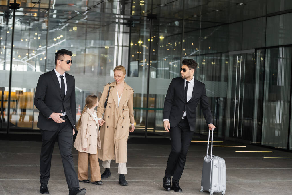 bodyguards protecting privacy of female clients, woman and girl walking out of hotel on blurred background, personal safety of rich family, men in sunglasses and suit with luggage   - Photo, Image