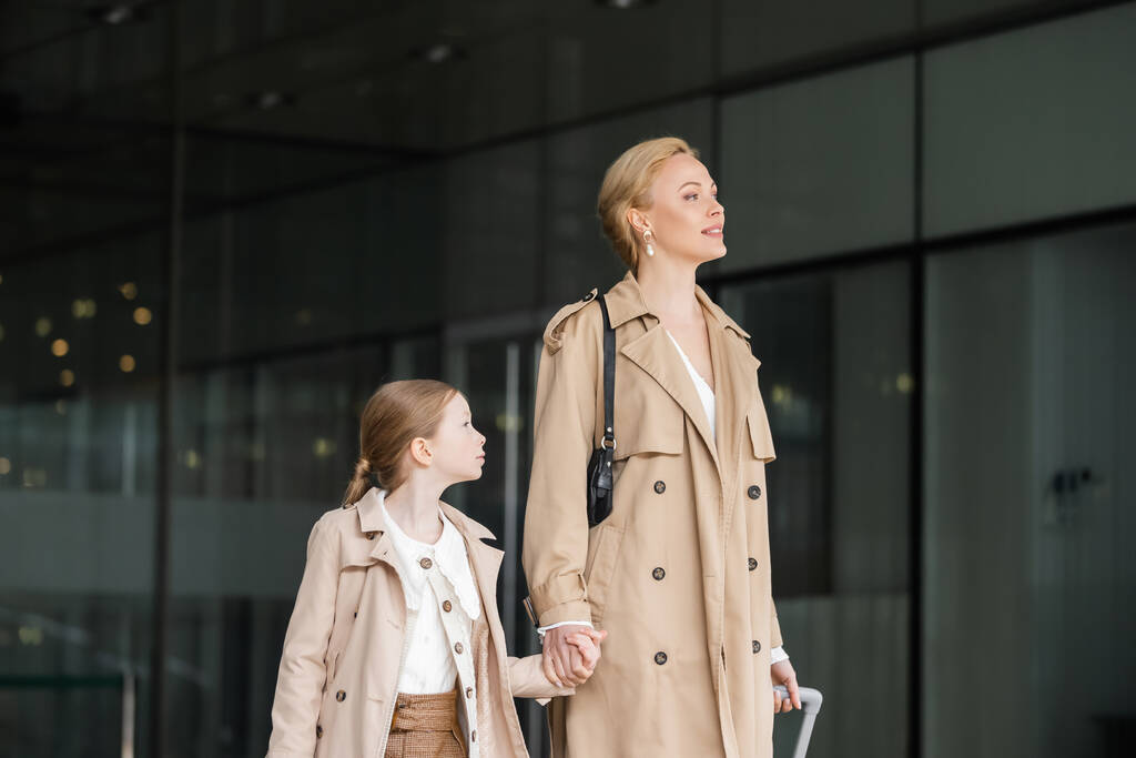 moderne mode, moeder dochter tijd, blonde vrouw met bagage vasthouden hand van tiener meisje terwijl lopen uit het hotel samen, slimme casual, beige trench jassen, buitenkleding, moderne ouderschap  - Foto, afbeelding