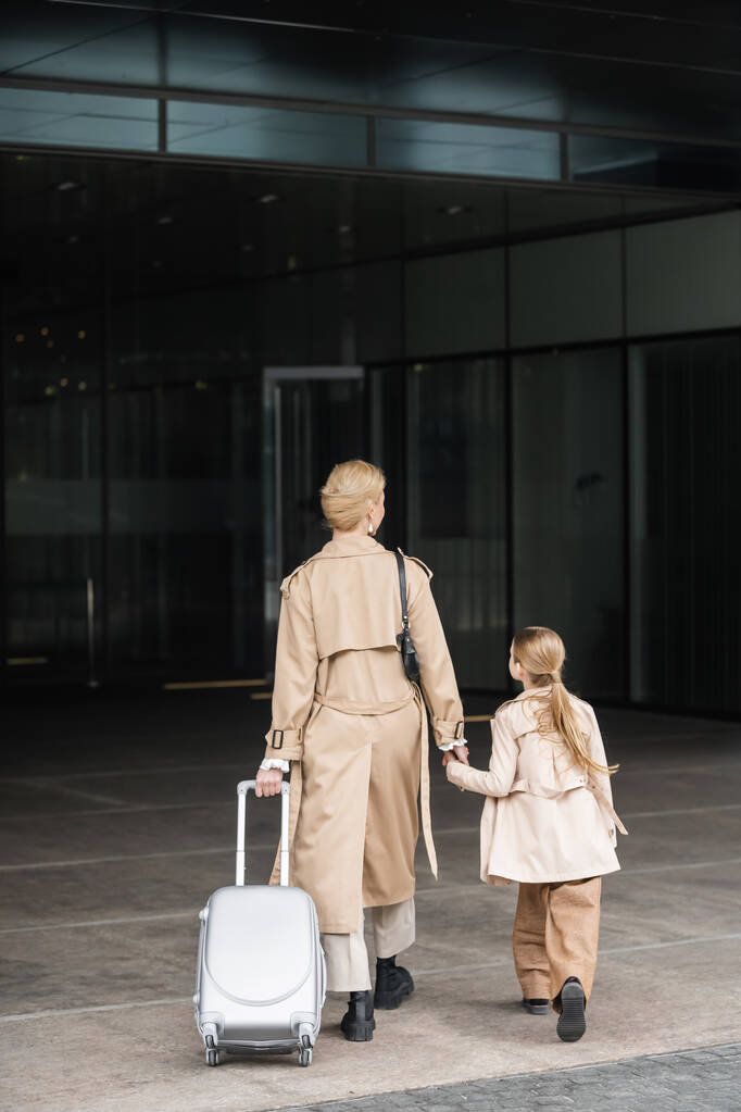 voyage en famille, mère et enfant, vue arrière de la femme blonde avec les bagages tenant la main de la fille tout en entrant dans l'hôtel ensemble, smart casual, beige trench coats, vêtements de dessus, smart casual  - Photo, image