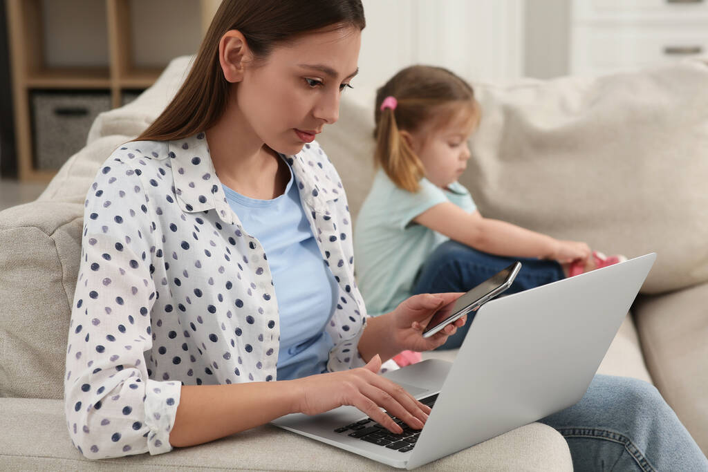 Femme avec ordinateur portable travaillant à distance à la maison. Mère et fille sur canapé dans le salon - Photo, image