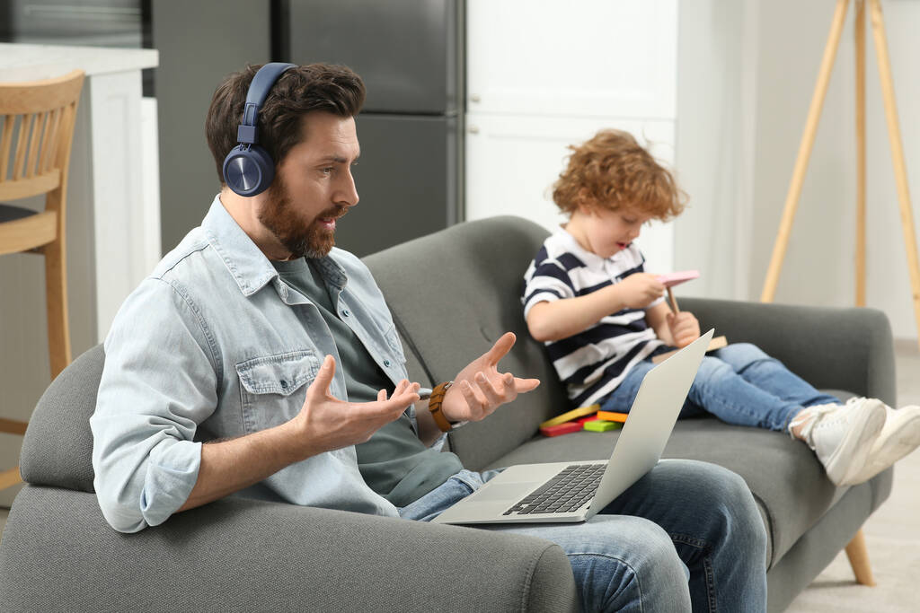 Father working remotely on laptop while his son playing with toys at home. Man having video chat with colleagues - Photo, Image