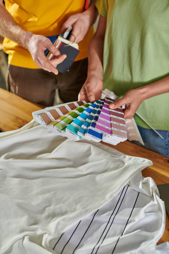 Καλλιεργημένη άποψη της νεαρής αφροαμερικανής βιοτεχνίας κρατώντας δείγματα χρωμάτων κοντά στο συνάδελφο δείχνοντας με το δάχτυλο και στέκεται κοντά σε ρούχα στο στούντιο εκτύπωσης, έννοια μικρή επιχειρηματική επιτυχία - Φωτογραφία, εικόνα