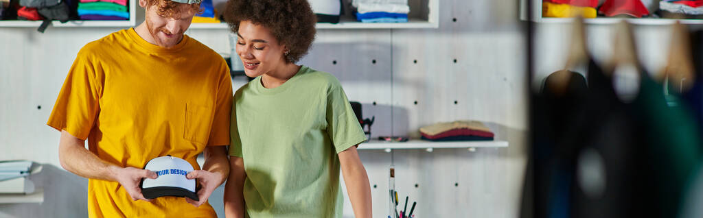 Jovens artesãos alegres e multiétnicos segurando snapback com camada de impressão enquanto trabalham em estúdio de impressão borrada em segundo plano, conceito de modelo de negócio sustentável, banner  - Foto, Imagem