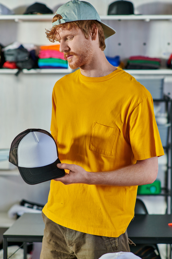Νεαρός κοκκινομάλλης τεχνίτης που κρατάει το στιγμιότυπο ενώ εργάζεται στο project και στέκεται στο θολό στούντιο εκτύπωσης στο παρασκήνιο, έννοια της ανθεκτικότητας στις μικρές επιχειρήσεις - Φωτογραφία, εικόνα