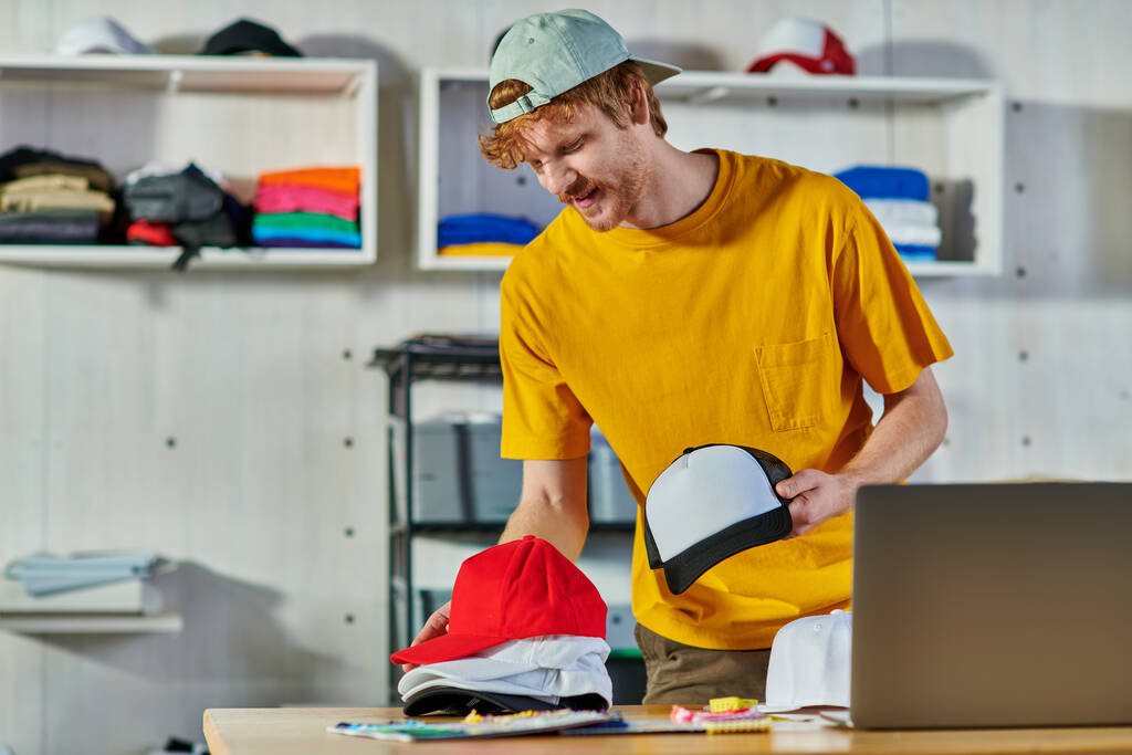 Cheerful young redhead craftsman holding snapbacks while working near cloth samples and blurred laptop on wooden table in print studio, small business resilience concept - Photo, Image