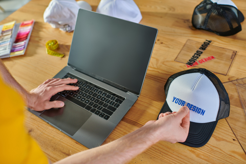 Widok projektanta korzystającego z laptopa z pustym ekranem i trzymającego warstwę drukującą w pobliżu migawki na rozmytym stole w pracowni druku, praktyczna koncepcja przedsiębiorczości  - Zdjęcie, obraz