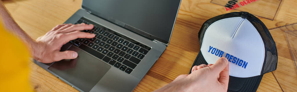 Высокоугольный вид молодого сумасшедшего человека, использующего ноутбук с экраном-блокнотом и держащего слой печати рядом со столиком в типографии, практическая предпринимательская концепция, баннер  - Фото, изображение