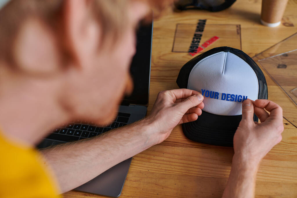 Blurred young designer holding printing layer near snapback while working on project near laptop on table in print studio, hands-on entrepreneurship concept, your design letters  - Photo, Image