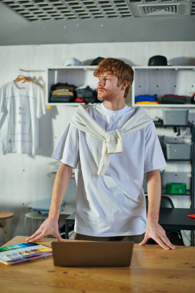 Young redhead craftsman in casual clothes looking away while standing near laptop and cloth samples on table in blurred print studio, self-made success concept  - Photo, Image