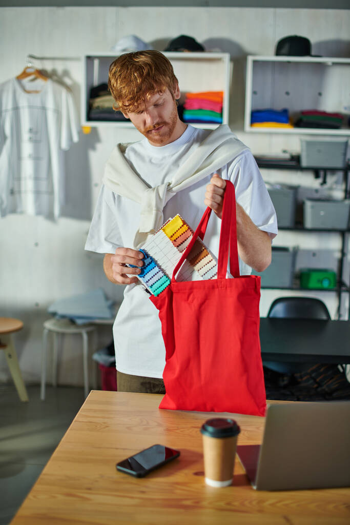Νεαρός κοκκινομάλλης τεχνίτης με casual ρούχα που κρατάει δείγματα υφάσματος κοντά στην τσάντα ώμου και συσκευές στο τραπέζι σε θολό στούντιο εκτύπωσης στο παρασκήνιο, αυτοδημιούργητη ιδέα επιτυχίας  - Φωτογραφία, εικόνα