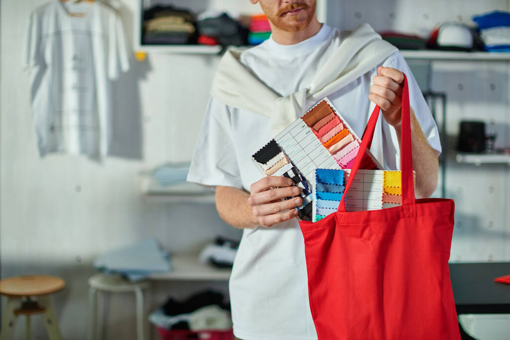 Cropped view of young craftsman in casual clothes putting cloth samples in shoulder bag while working in blurred print studio at background, self-made success concept  - Photo, Image