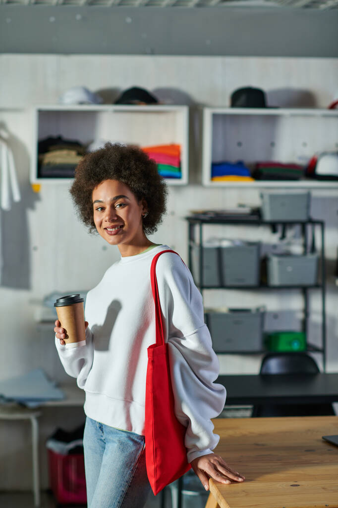 Usmívající se mladá afro-americká řemeslnice s taškou na rameno, která drží kávu a dívá se na kameru u stolu v rozmazaném tiskovém studiu, nadšená podnikatelka pracující v dílně - Fotografie, Obrázek