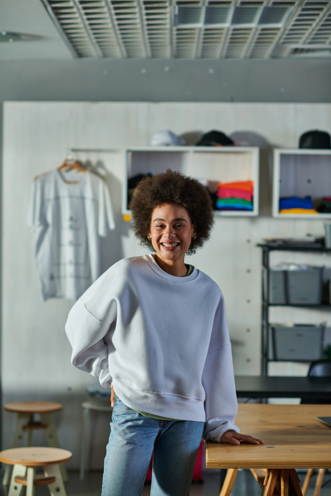 Веселая и уверенная в себе африканская ремесленница в толстовке и джинсах, смотрящая в камеру, стоя возле рабочего стола в типографии, восторженный владелец бизнеса, работающий в мастерской - Фото, изображение