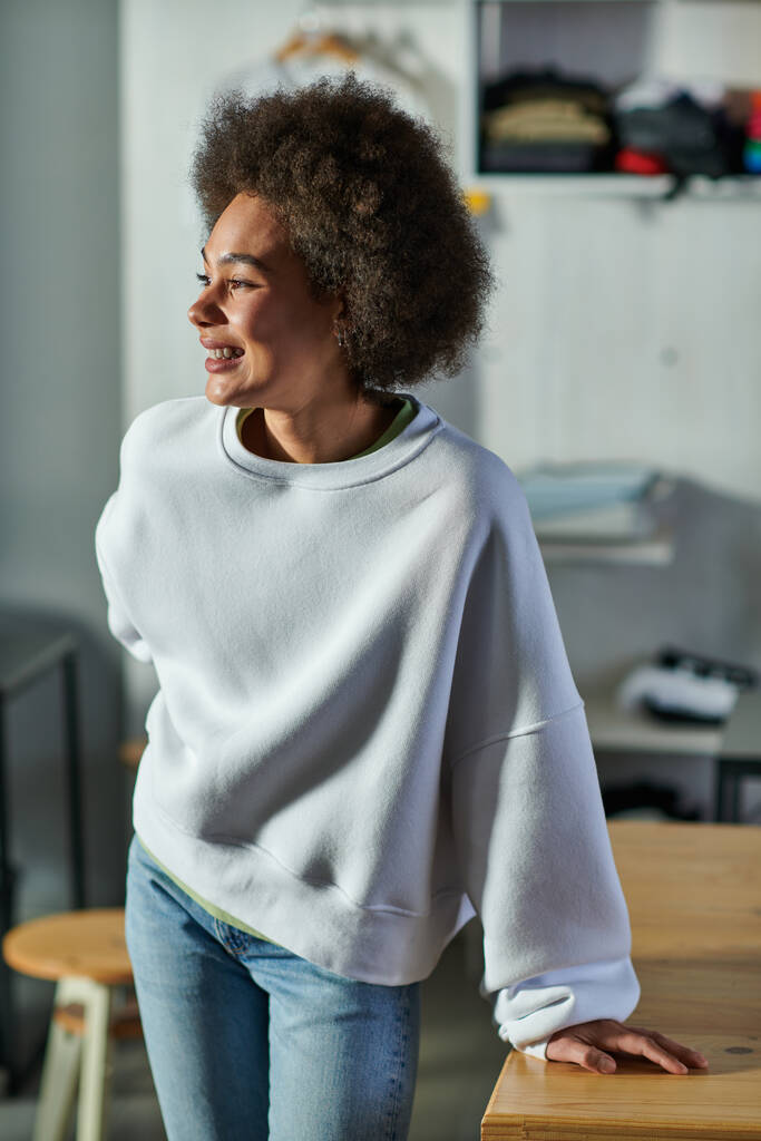 Portrait of cheerful young african american craftswoman in sweatshirt and jeans looking away while standing near working table in blurred print studio, enthusiastic business owner working in workshop - Photo, Image